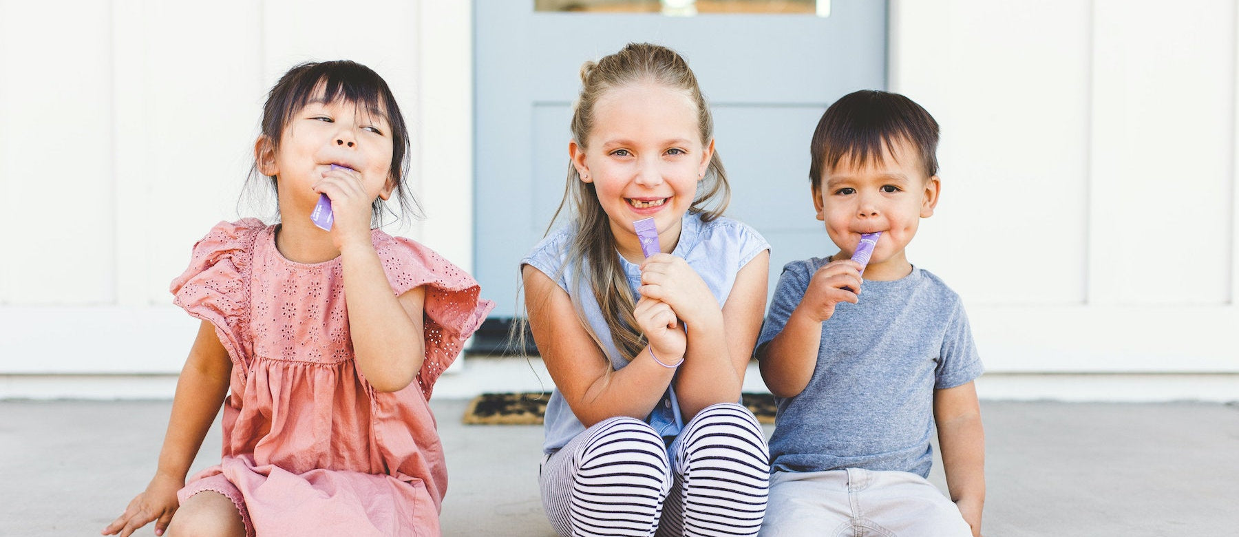 3 young children sitting on top step of porch holding stick packs of immune booster to mouth